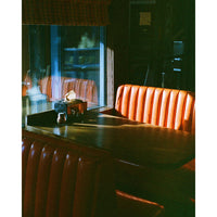 A dimly lit restaurant booth with orange-brown pleated upholstery and a dark wooden table, photographed in moody low light with reflections on a window.
