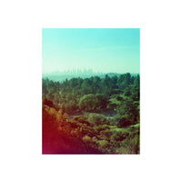 A vertical photographic art print depicting a scenic landscape with a distant city skyline on the horizon, a line of green trees, and sunlit hills in the foreground, presented with a white border.