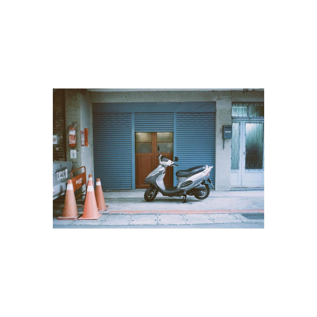 A silver and black scooter parked on a sidewalk in front of a closed blue metal shutter door, with orange traffic cones on the left and a white door on the right.
