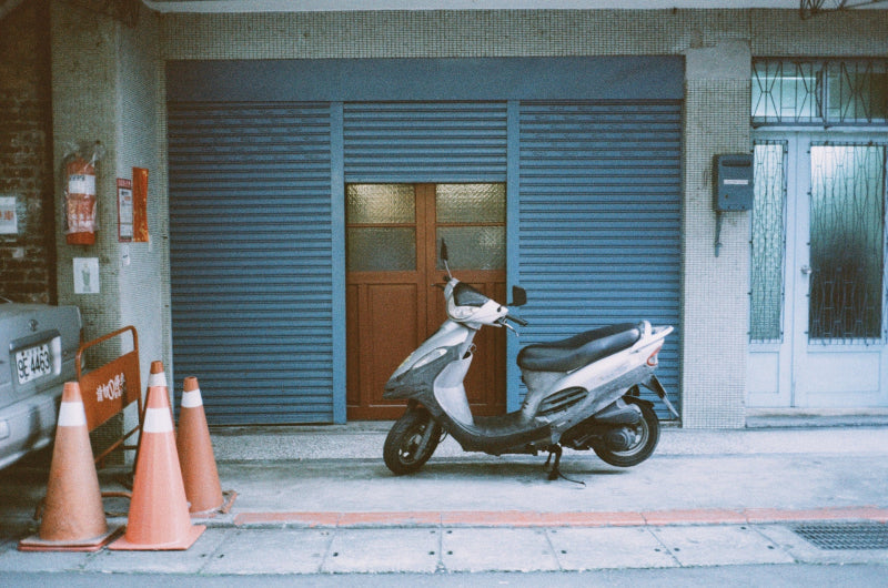 A silver and black scooter parked on a sidewalk in front of a closed blue metal shutter door, with orange traffic cones on the left and a white door on the right.