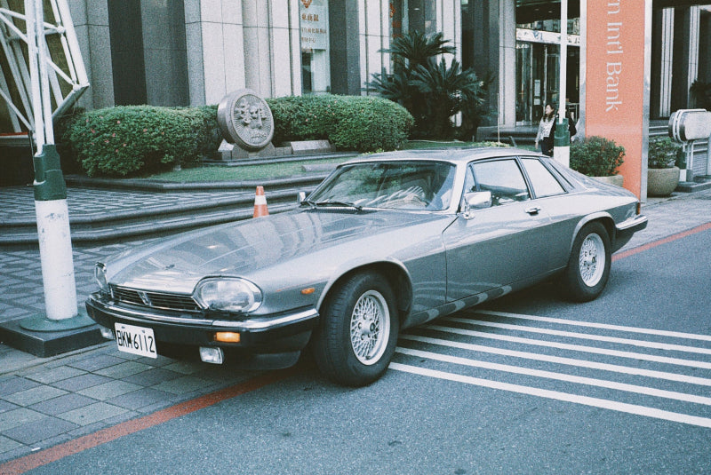 A silver Jaguar S-Type coupe parked on a city street in front of a modern building, with a Japanese license plate visible.