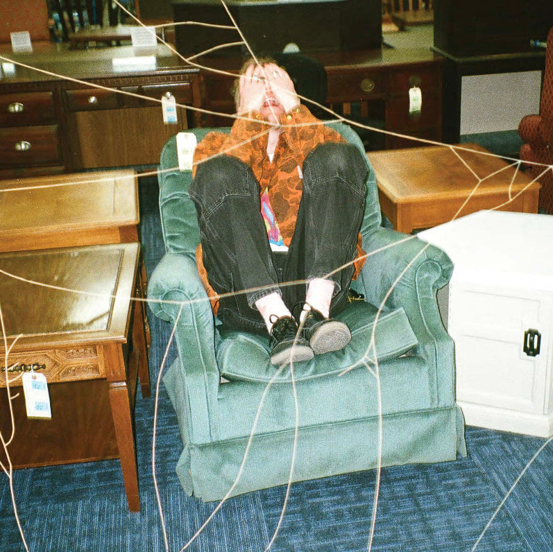A person reclining in a dusty green armchair inside a furniture store, surrounded by other chairs, wooden tables, and a white storage cabinet. The photo has a web-like overlay pattern across the frame.