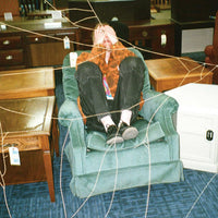 A person reclining in a dusty green armchair inside a furniture store, surrounded by other chairs, wooden tables, and a white storage cabinet. The photo has a web-like overlay pattern across the frame.