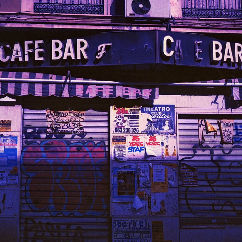 A street-level view of a closed cafe bar storefront with graffiti-covered metal shutters, posters, and a dark awning. The scene has a strong purple cast and includes signage reading 'CAFE BAR' across the top.