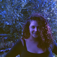 A woman with long, curly dark hair wearing a black top, photographed outdoors at night with blue-toned lighting against a background of trees.