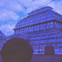 A blue-toned photograph of a large glass conservatory building with arched metal framing, set against a cloudy sky, with trimmed hedges in the foreground.