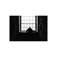 A black and white photograph of a person sitting in silhouette at a piano in a dimly lit stone room, facing a large multi-paned window.