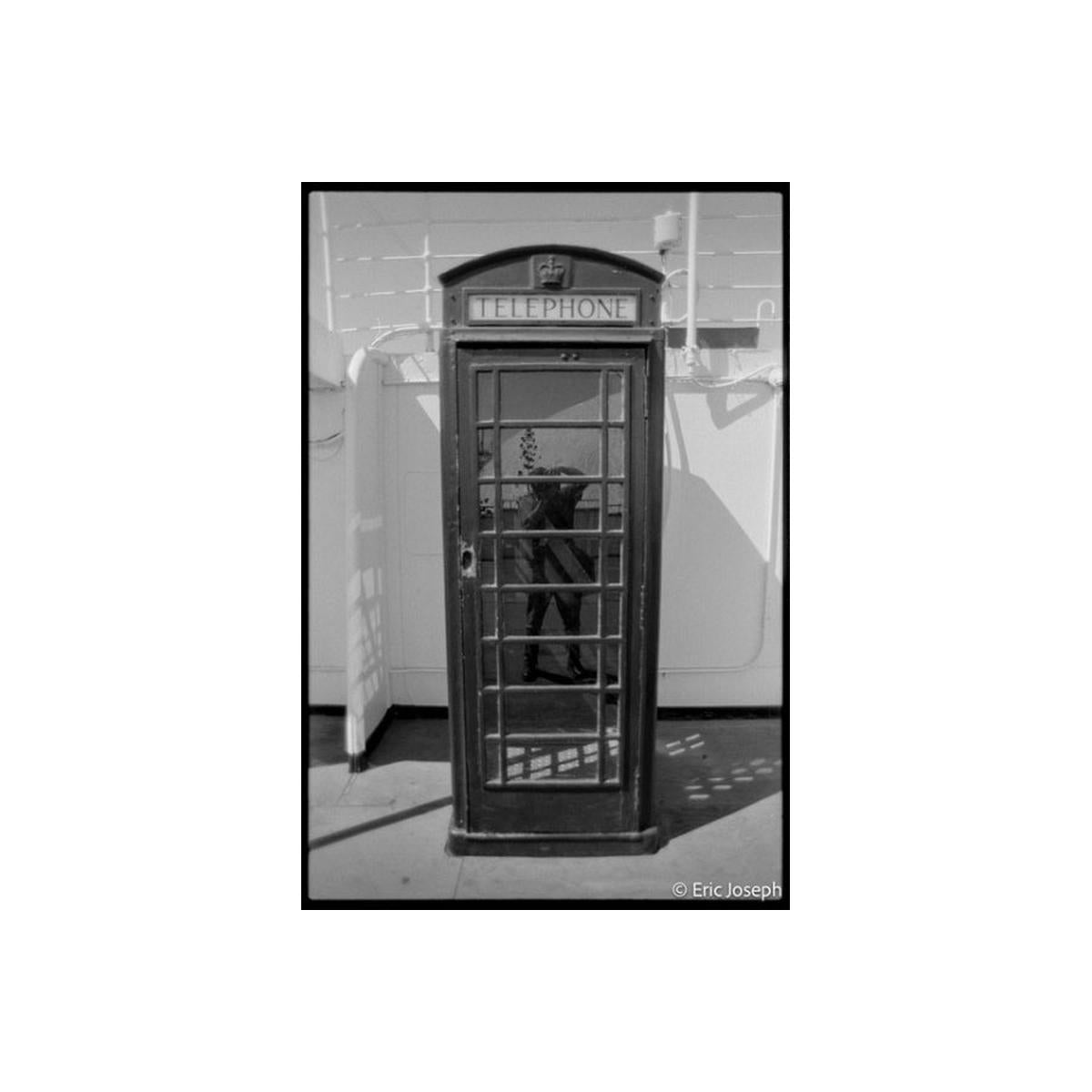 A black and white photograph of a classic British telephone booth, with a reflection of a person visible in the glass door. The words 'TELEPHONE' are printed on the top sign.