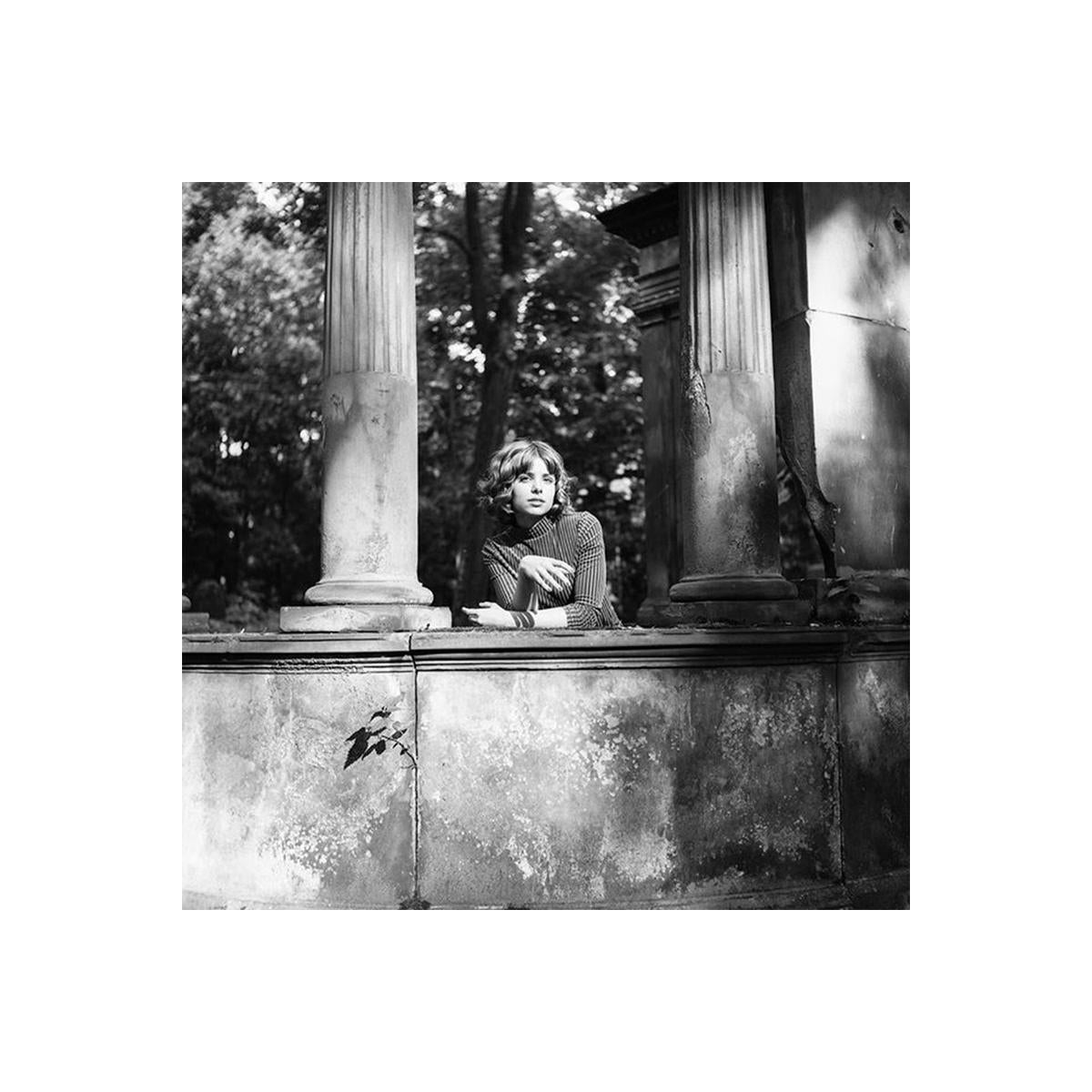 Black and white medium-format portrait of a woman leaning against stone pillars in a garden, shot on Rollei RPX 100 film.