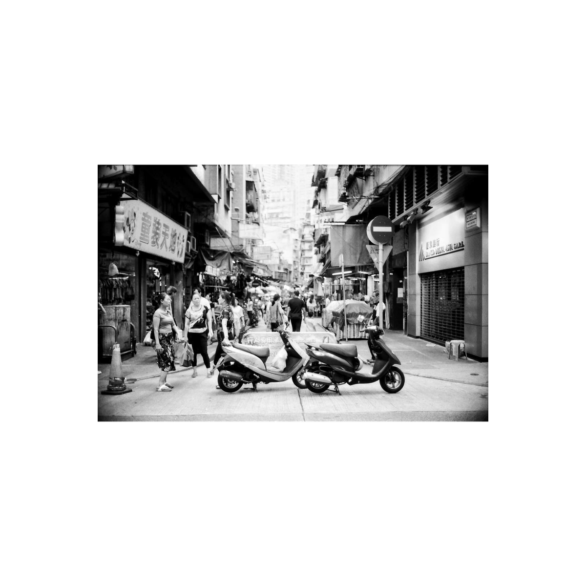 Black and white street scene featuring two parked scooters on a narrow city alleyway with pedestrians and storefronts.