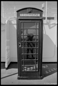 A black and white photograph of a classic British telephone booth, with a reflection of a person visible in the glass door. The words 'TELEPHONE' are printed on the top sign.