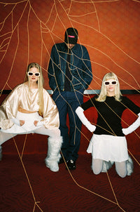 Three models pose in a stylized studio setup with a textured orange-brown wall, gold web-like lines, and a dark patterned floor. One model wears a shiny cream satin top and white pants, seated on a bench.
