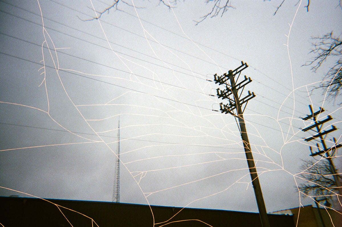 An artistic photograph of utility poles and power lines against a cloudy gray sky, overlaid with long exposure light trails in white and gold.