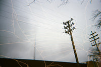 An artistic photograph of utility poles and power lines against a cloudy gray sky, overlaid with long exposure light trails in white and gold.