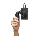 Hand holding a black Sony RX100 VII camera with a ZEISS lens and attached shooting grip against a white background.