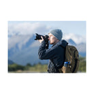 Photographer using a black Sony a7R VI mirrorless camera with a lens outdoors against a blurred mountain landscape.