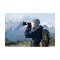 Photographer using a black Sony a7R VI mirrorless camera with a lens outdoors against a blurred mountain landscape.
