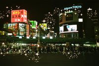 A nighttime city scene with brightly lit billboards and signs, including visible text such as AlpenTokyo and Japanese signage, with a crowd of people crossing a street in the foreground. The image has decorative sparkle/star overlays.
