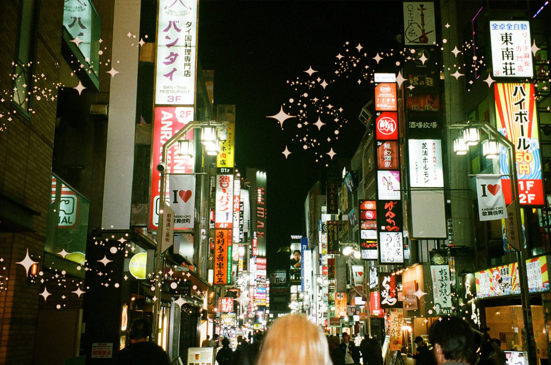 A nighttime street scene in a Japanese city with glowing multicolored neon signs, storefront signs in Japanese, and many blurred pedestrians walking down the street. The image has a warm vintage filter with soft focus and decorative starburst overlays.