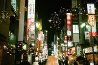 A nighttime street scene in a Japanese city with glowing multicolored neon signs, storefront signs in Japanese, and many blurred pedestrians walking down the street. The image has a warm vintage filter with soft focus and decorative starburst overlays.