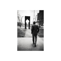 A black and white photograph of a man in a suit walking away from the camera toward a modern abstract metal sculpture in an urban park setting.