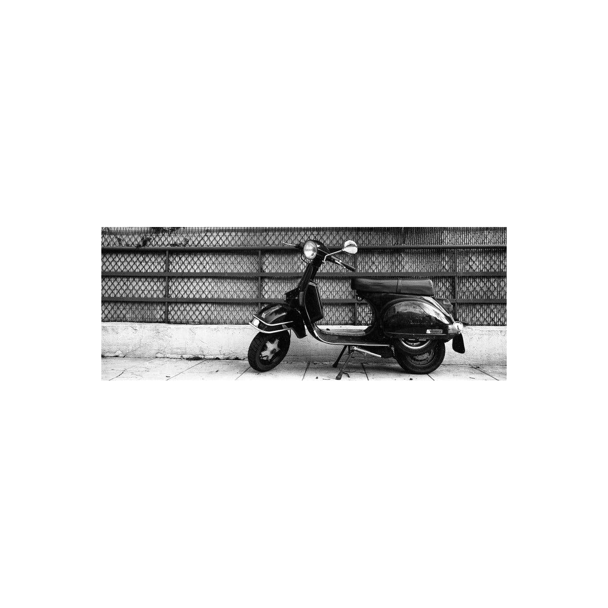 A black vintage-style scooter parked on a sidewalk in front of a chain-link fence, shown in a monochrome photograph.