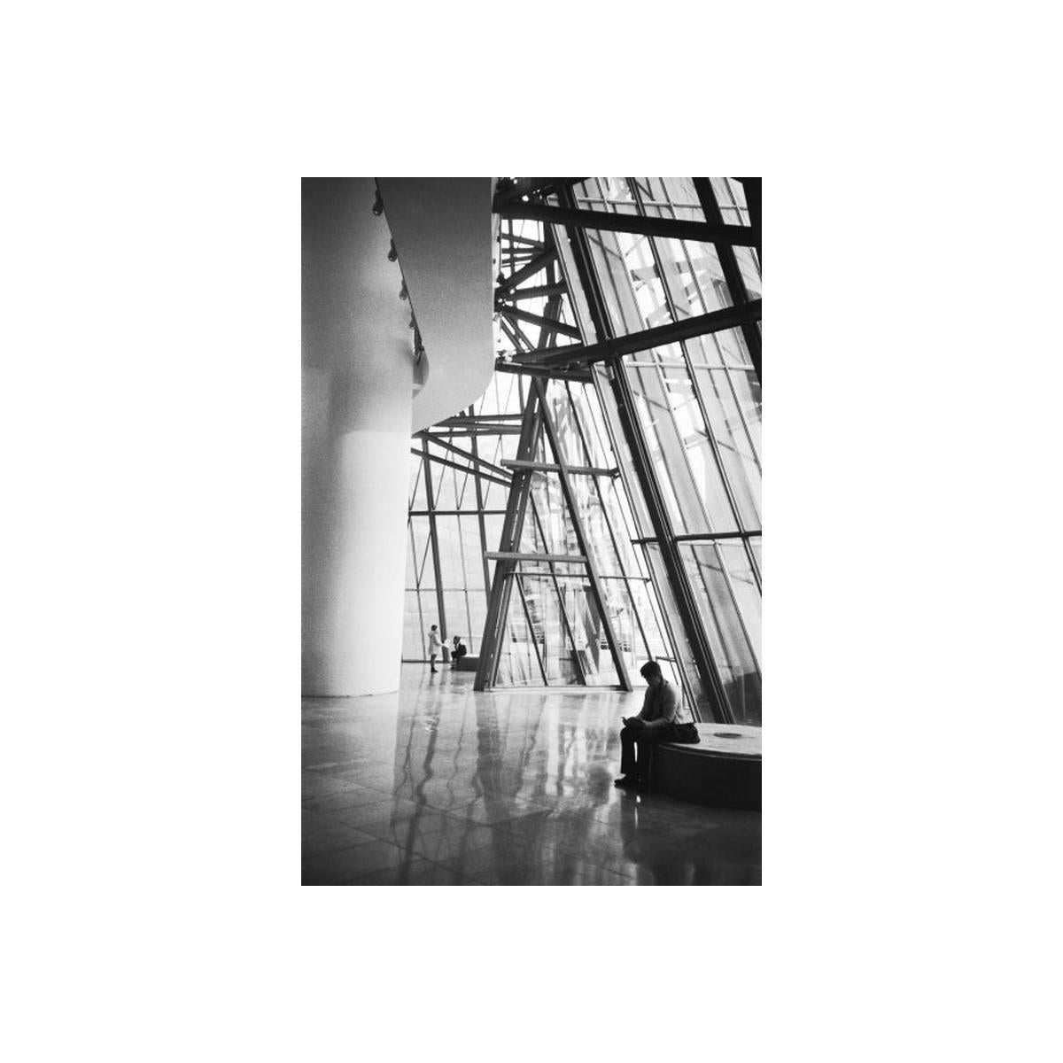Black and white architectural photograph of a modern glass-walled interior space, with a seated person visible near the lower right and large structural columns and beams throughout the scene.