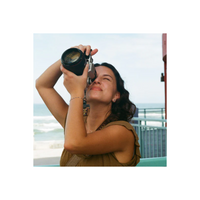 A woman holding a vintage film camera up to her eye, looking through the viewfinder. She is wearing a brown sleeveless top, with a beach and ocean railing in the background.