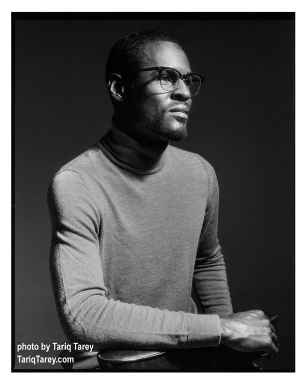 A black and white portrait photograph of a man wearing thick-rimmed glasses and a gray turtleneck sweater, seated against a dark studio background. White text in the lower-left corner reads 'photo by Tariq Tarey'.