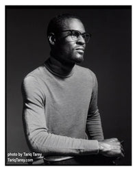 A black and white portrait photograph of a man wearing thick-rimmed glasses and a gray turtleneck sweater, seated against a dark studio background. White text in the lower-left corner reads 'photo by Tariq Tarey'.