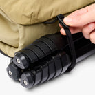 Person securing a black tripod to a beige bag with a strap.
