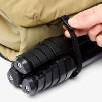 Person securing a black tripod to a beige bag with a strap.
