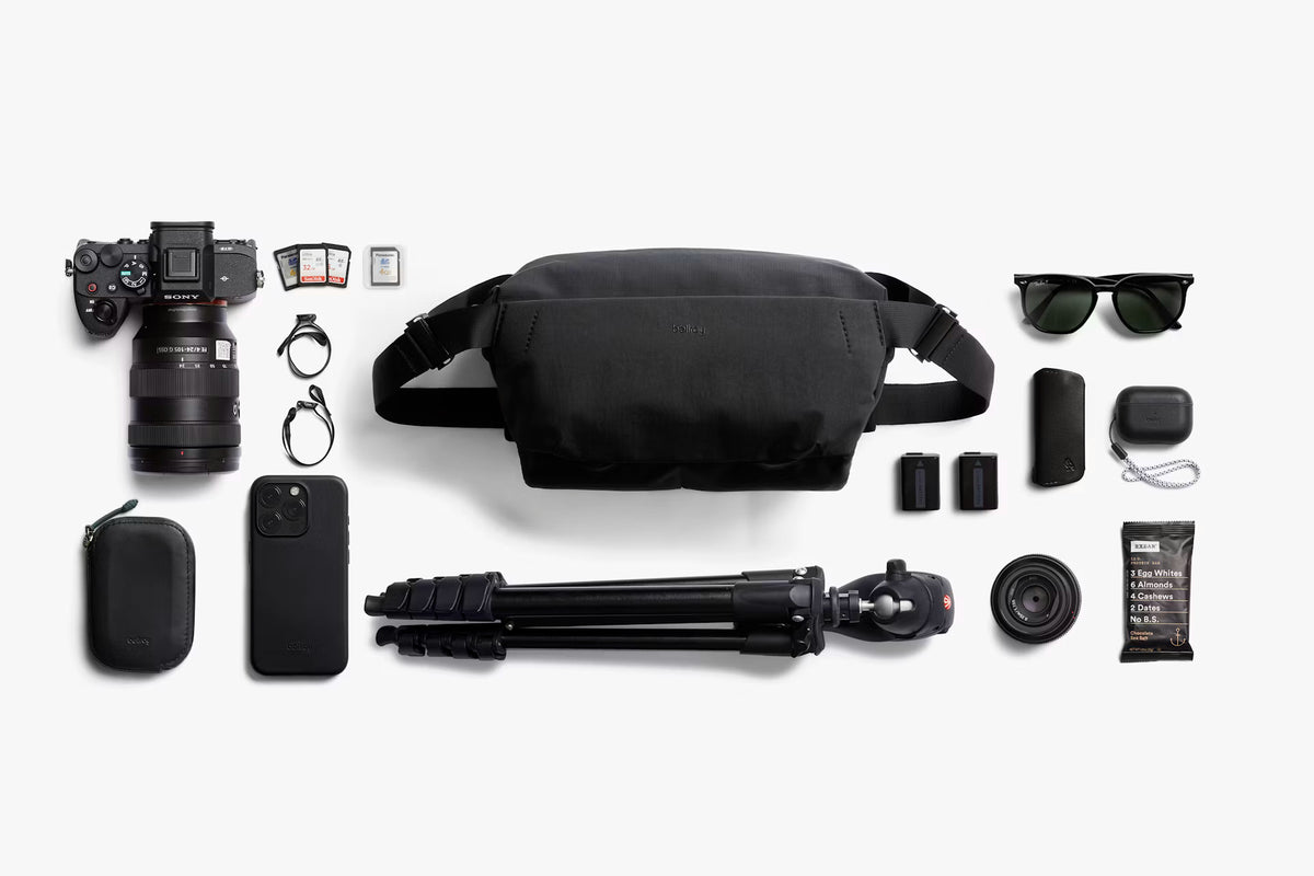 A black camera-focused lifestyle flat lay featuring a black waist bag, a DSLR camera, a tripod, sunglasses, a smartphone, and various accessories arranged neatly on a light gray background.