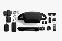 A black camera-focused lifestyle flat lay featuring a black waist bag, a DSLR camera, a tripod, sunglasses, a smartphone, and various accessories arranged neatly on a light gray background.