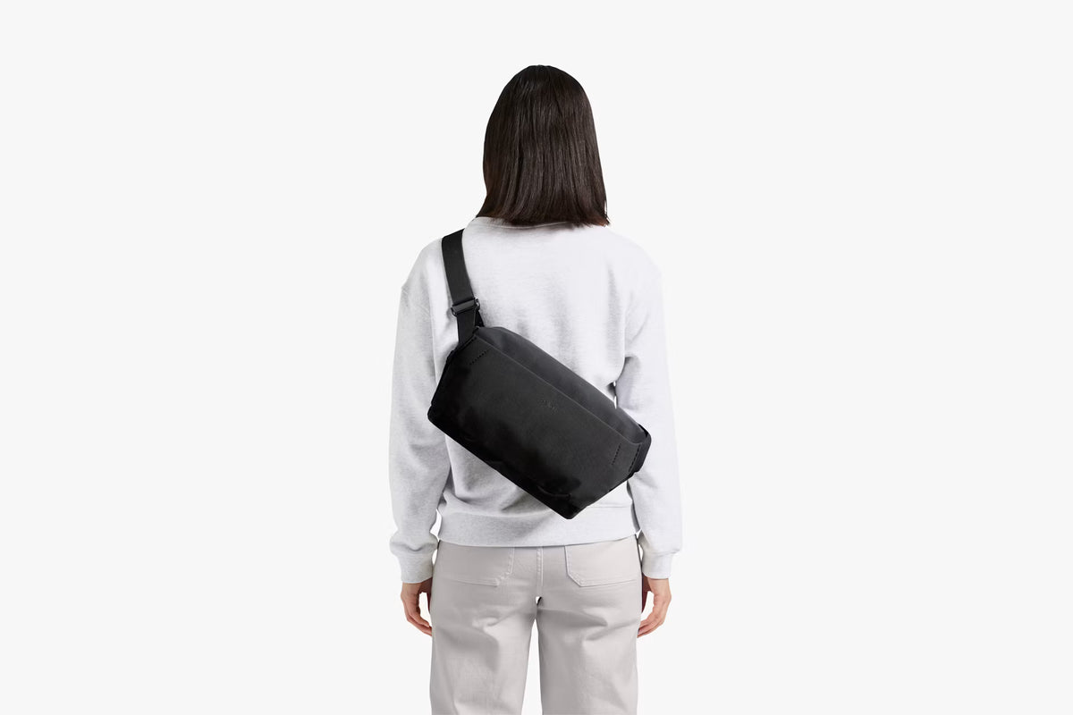 A woman viewed from behind wearing a light gray sweatshirt and light gray pants, carrying a black rectangular crossbody bag across her back against a plain white background.