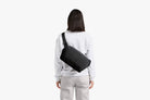 A woman viewed from behind wearing a light gray sweatshirt and light gray pants, carrying a black rectangular crossbody bag across her back against a plain white background.