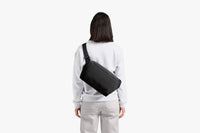 A woman viewed from behind wearing a light gray sweatshirt and light gray pants, carrying a black rectangular crossbody bag across her back against a plain white background.