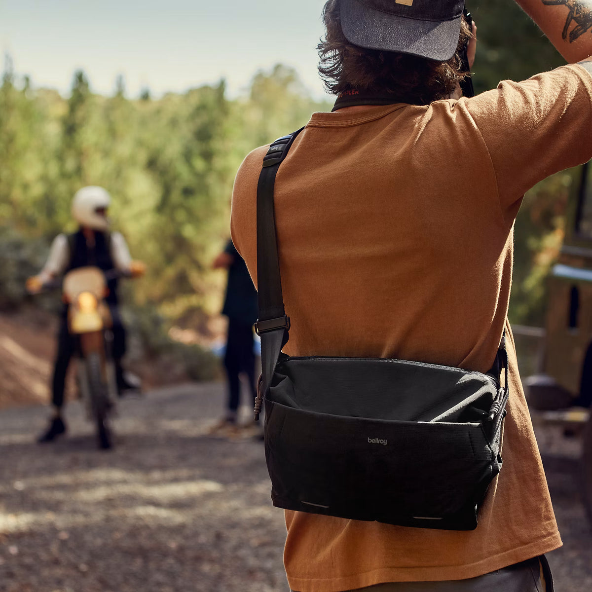 Person with a black bag on a dirt road with another person on a motorcycle in the background.