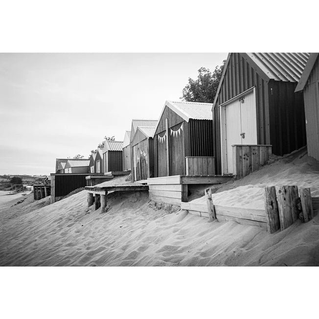 Black and white photograph of a row of wooden beach huts with corrugated metal roofs situated on a sandy dune, with a pale sky in the background.