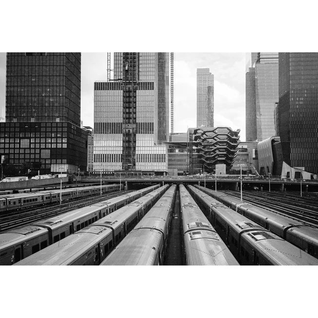 Black and white photograph of a city train station with multiple trains on tracks leading toward modern high-rise buildings.