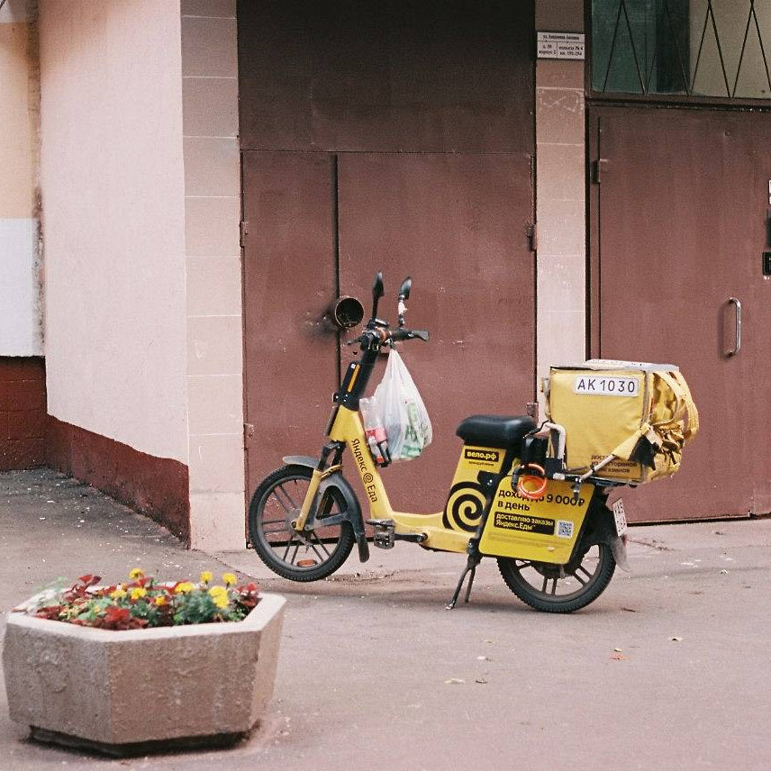 A yellow electric scooter with a delivery box on the back is parked on a sidewalk in front of a building with brown doors and a planter box in the foreground.