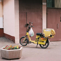 A yellow electric scooter with a delivery box on the back is parked on a sidewalk in front of a building with brown doors and a planter box in the foreground.
