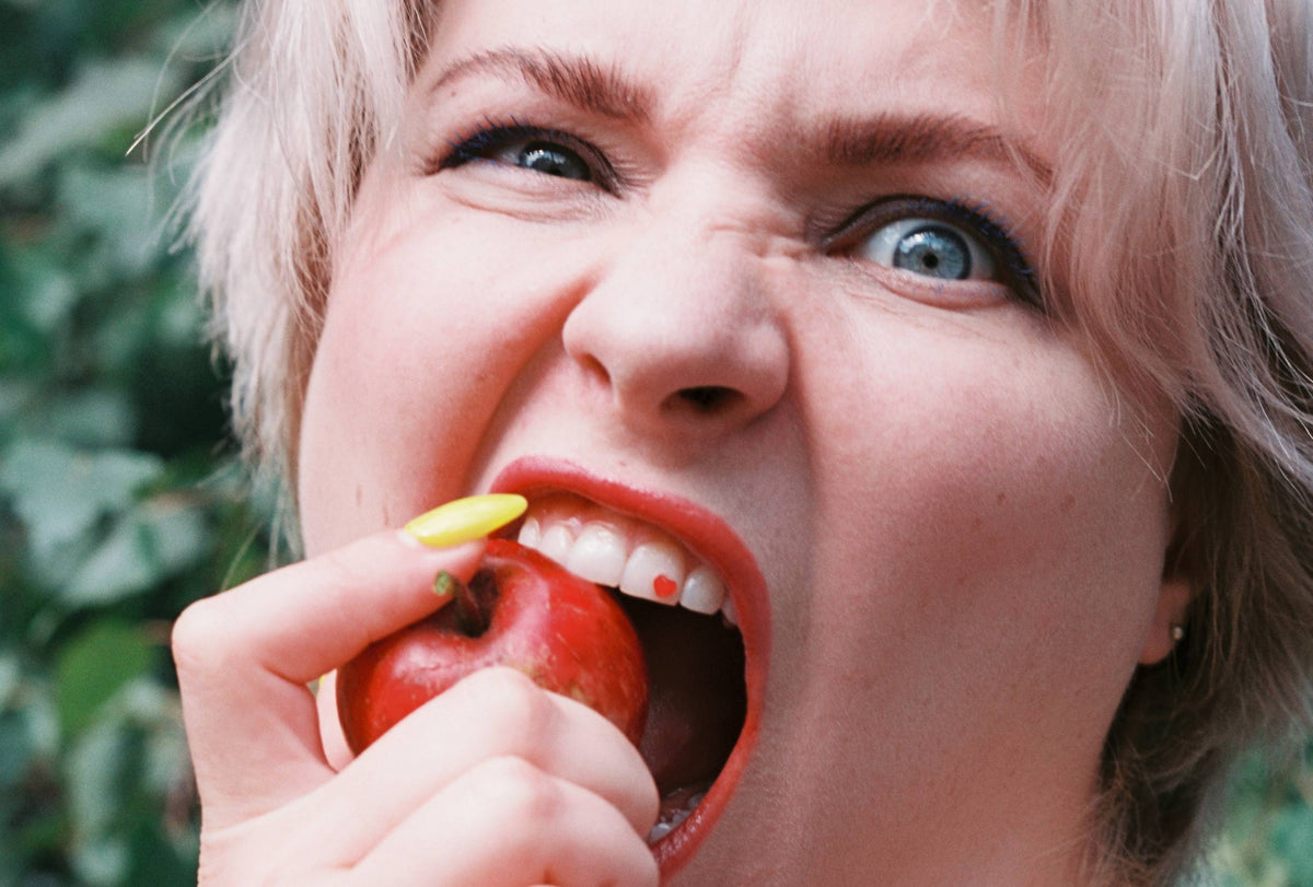 A close-up lifestyle shot of a woman with short blonde hair biting into a red apple, holding a yellow object near her mouth, with a blurred green outdoor background.