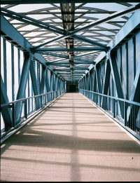 A perspective view down a covered bridge walkway with a blue steel truss structure and a beige floor, framed by repeating blue beams.