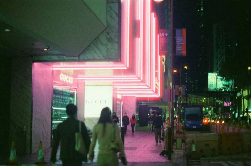 A nighttime street scene featuring a brightly lit Gucci storefront with pink and white neon-like lighting, with pedestrians walking in front of the entrance.