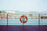 An orange life preserver with white stripes hangs on a metal railing overlooking a harbor with boats, water, and distant mountains under a pale blue sky.