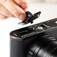 A close-up of a hand removing a black camera lens protector cap from the side of a black compact camera.