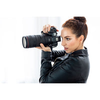 A woman with her hair in a bun wearing a black leather jacket and holding a black DSLR camera with a large lens, set against a bright white background.