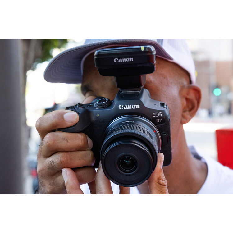 A close-up lifestyle shot of a person holding a Canon EOS R7 mirrorless camera with an attached lens and a mounted flash, wearing a white cap and white shirt against a blurred outdoor street background.