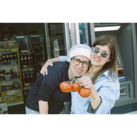 A smiling couple posing together in front of a convenience store. The man wears a white beanie, glasses, and a black shirt, while the woman wears a light blue button-up shirt and sunglasses.